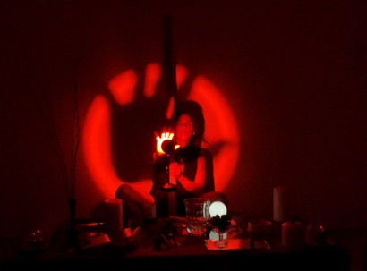 Paola is crouched between a series of found objects which are placed on an altar made of rock. She is illuminated with a red spotlight and has one hand lifted creating a shadow.